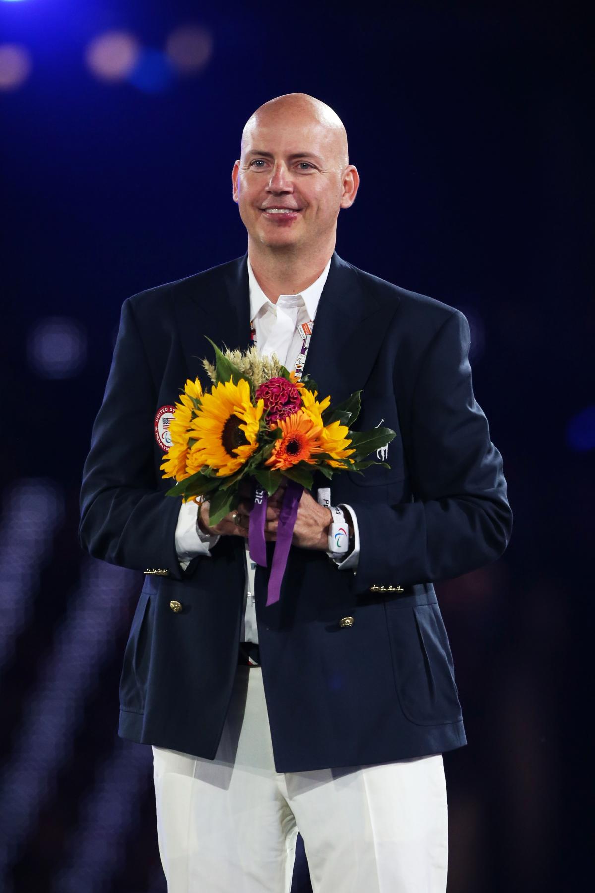 A picture of a smartly dress man, with light trousers and dark jacket holding a bunch of flowers.