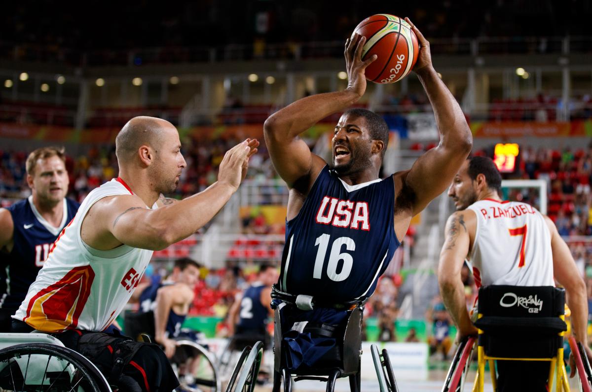 Man in wheelchair holds a basketball in the air away from his defender