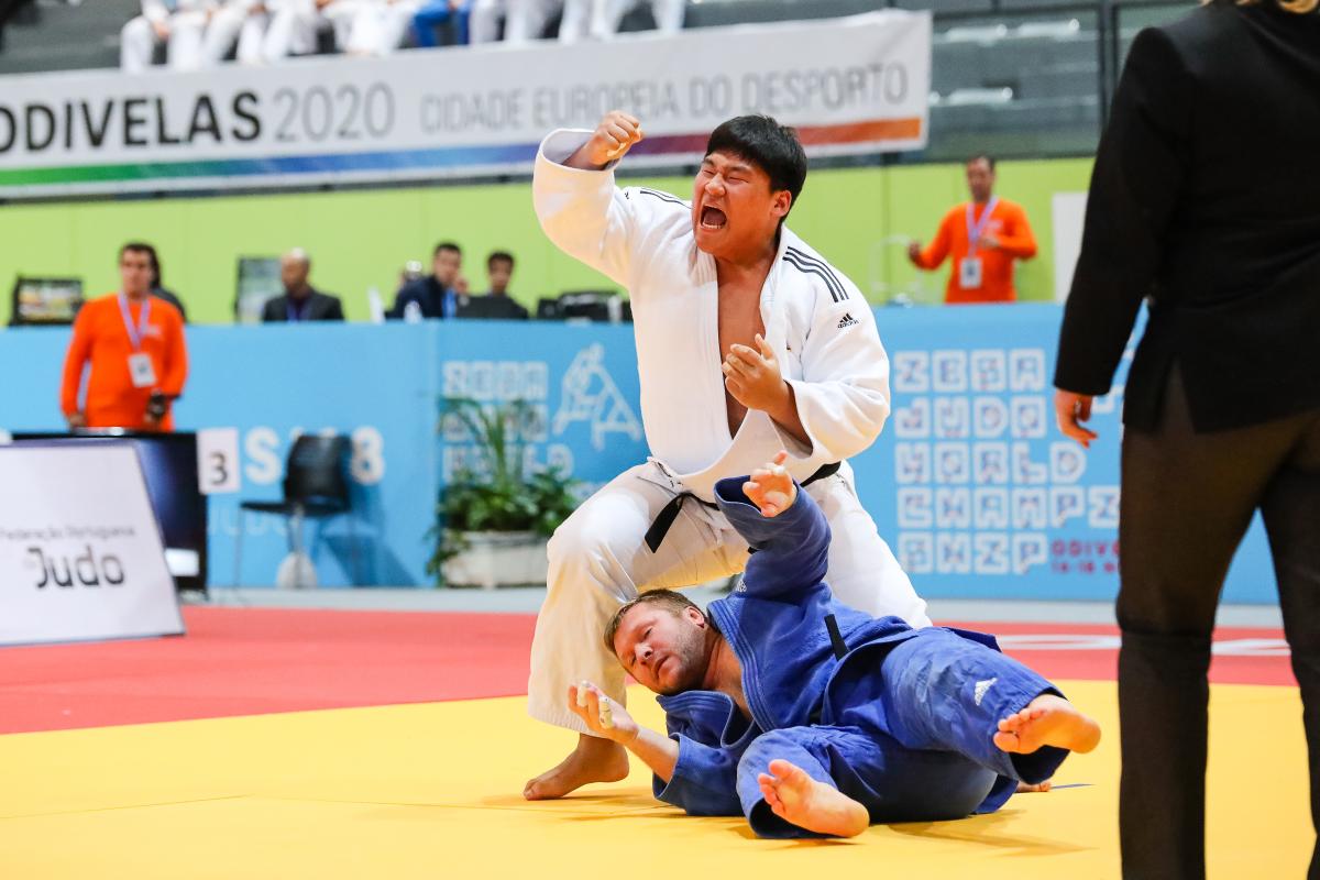 A man in a white kimono celebrates while other man in blue kimono lies on the mat