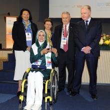 Iranian archer Zahra Nemati seated in a wheelchair holding an award surrounded by people.