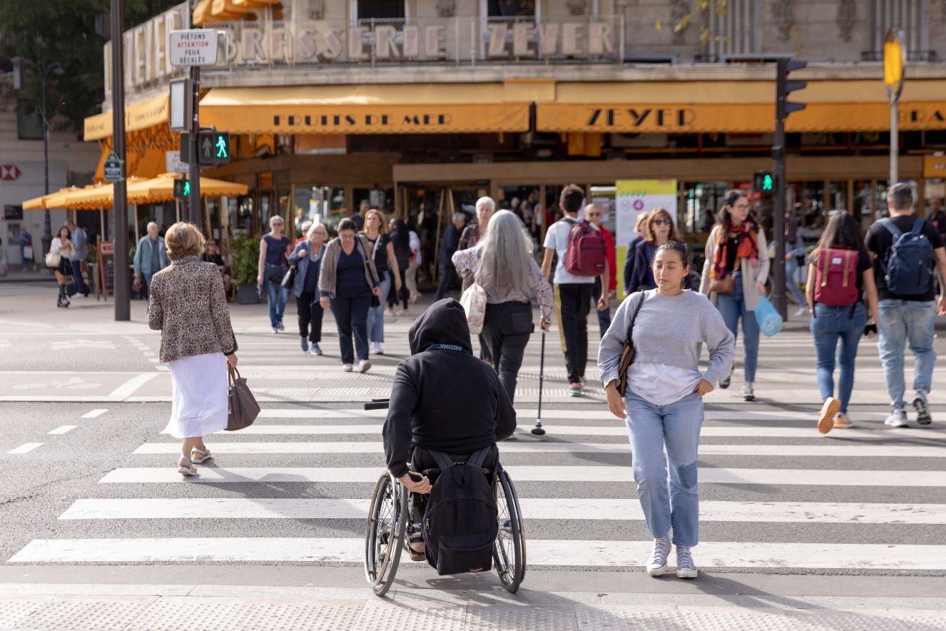 Many people cross a sidewalk in Paris. There are people in a wheelchair and people using canes.