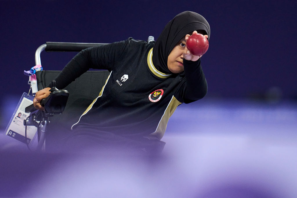 A woman in a wheelchair holds up a boccia ball and prepares to throws it