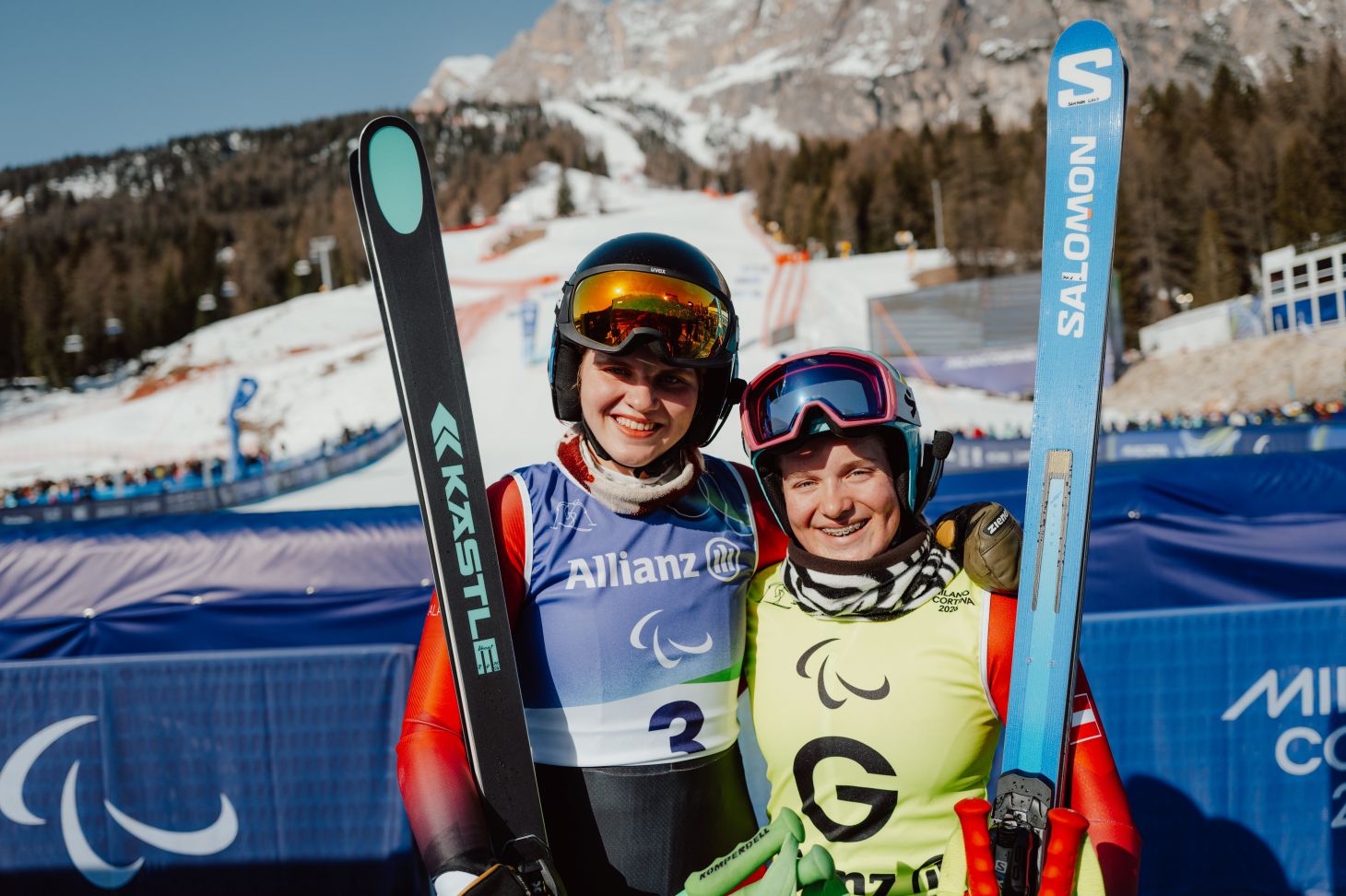 Two female skiers - one Para athlete and her guide - pose for a camera in front of the slopes