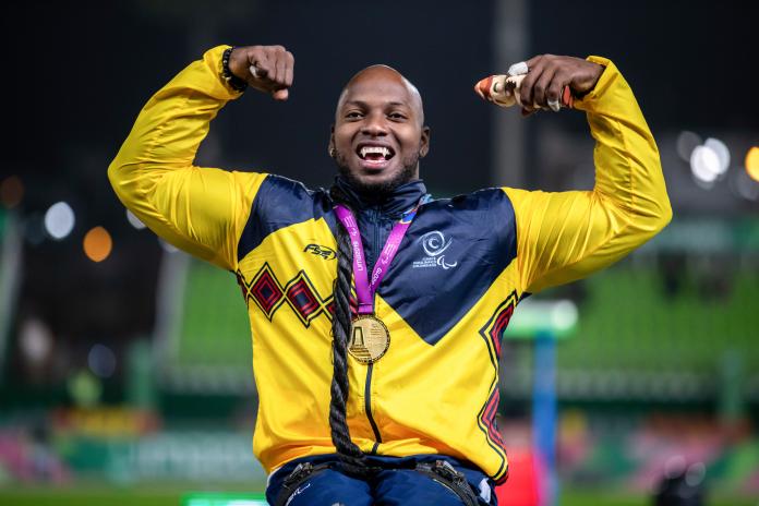 Un atleta masculino en silla de ruedas con una medalla de oro alrededor de su cuello levanta las manos