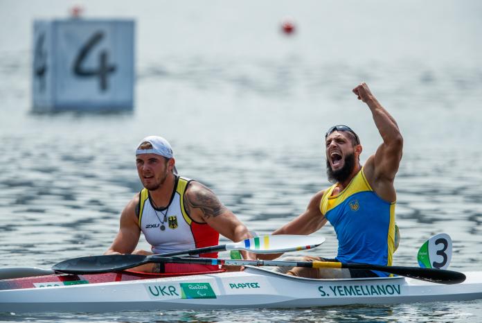 two male Para canoeists side by side in their boats with one punching the air in celebration