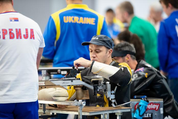 A man loading his rifle during a shooting competition with other competitors and a man standing next to him