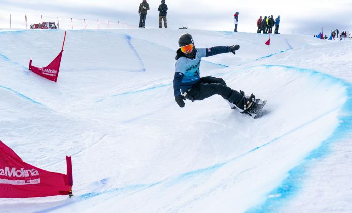 A male Para snowboarder competing being observed by eight people in the background