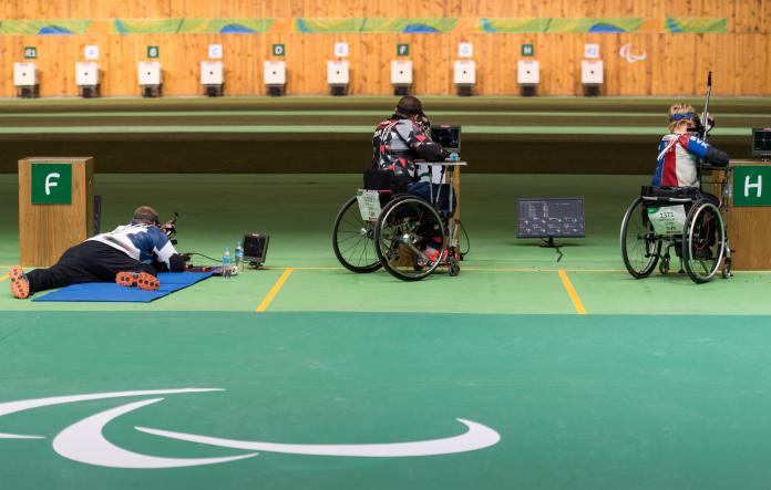 A man on the floor and two persons in wheelchairs with rifles aiming at targets in a shooting Para sport competition