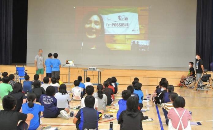 School children in a gym looking at a screen with a woman speaking via video call