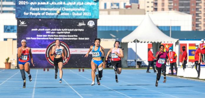 A group of five women with prosthetic legs running on a blue athletics track