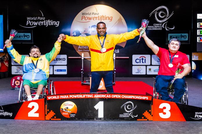 A man standing in the centre of a podium holding the hands of two other men in wheelchairs