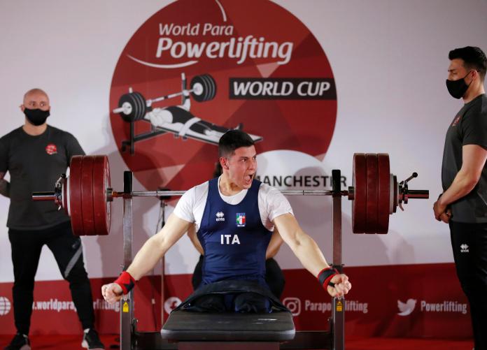 A men without legs on a bench press celebrating observed by two other men standing during a Para powerlifting competition