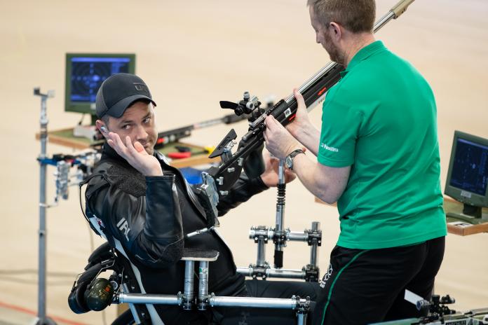 Male shooting athlete waves at crowd as his assistant helps