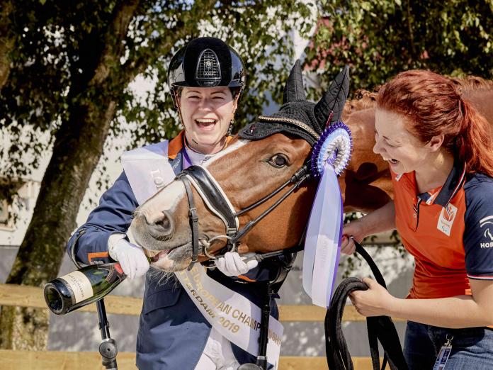 Woman smiling with her horse who is biting a champagne bottle