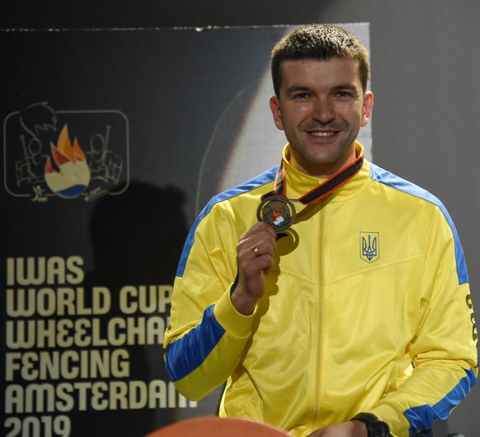 Ukrainian wheelchair fencer Andrii Demchuk smiles and poses to the camera with his gold medal