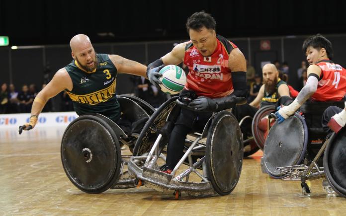 Japanese wheelchair rugby player tries to advance in posession with the ball while Australian player tries to prevent it