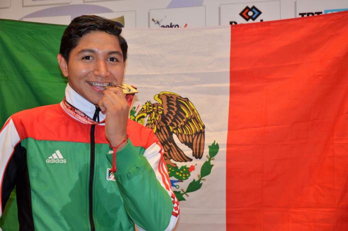 Male Mexican taekwondo athlete smiles biting medal in front of Mexican flag