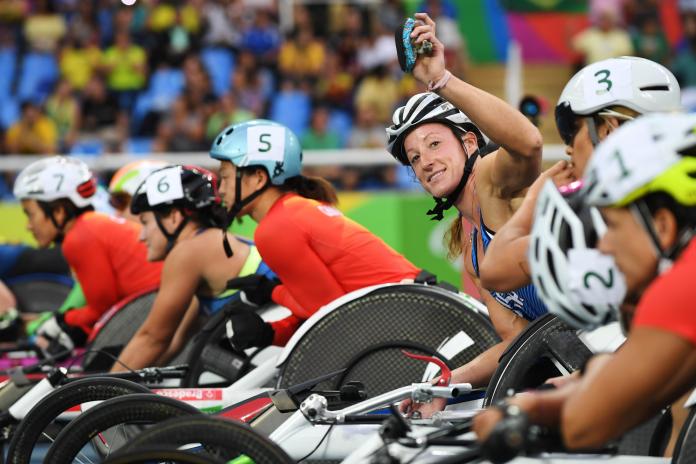 A woman in a racing wheelchair waving to the camera surrounded by other six female wheelchair racers