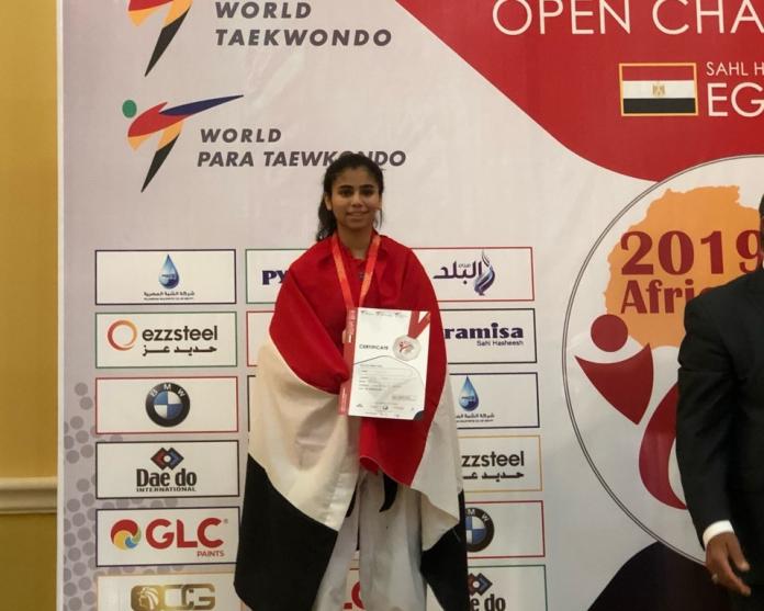 Egyptian teenage taekwondo athlete smiles on podium with flag around her