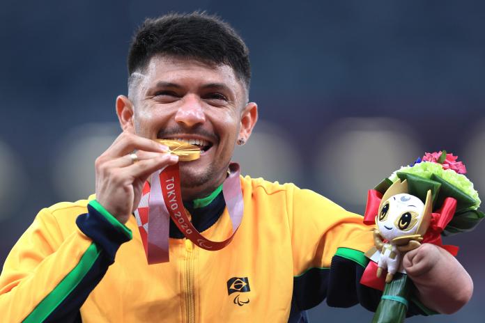 A man bites the gold medal with a smile on his face