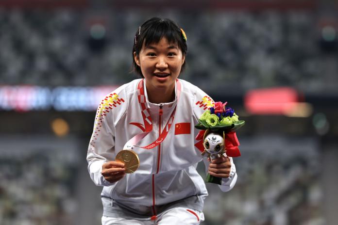 Xiaoyan Wen crouches on the podium displaying gold medal and mascot