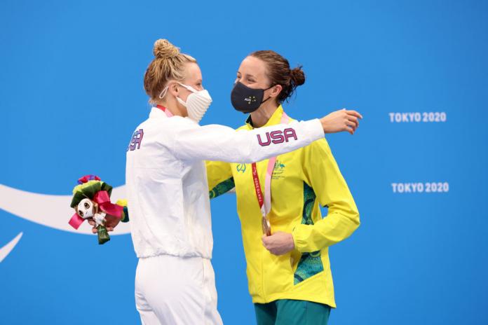 Two women hugging in front of a backdrop showing the Paralympic Agitos in white