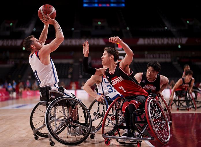 Man in wheelchair shoots basketball while defender reaches out to block the shot