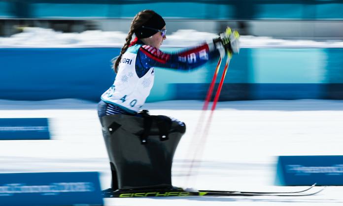 A woman in a sit ski competition pushing herself forward with two sticks.