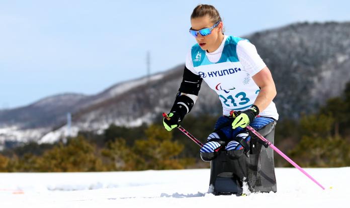 A woman with goggles pushing herself forward with two sticks in a sit ski competition on a sunny day.