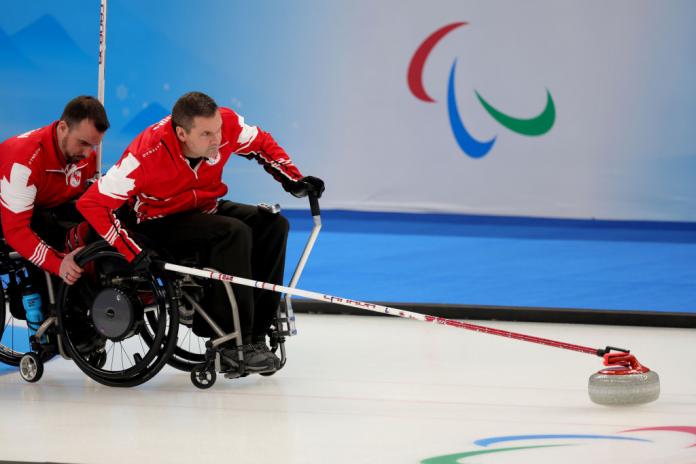 Mark ideson of Canada is held by a teammate as he goes to release the curling stone at Beijing 2022