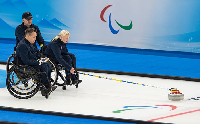 A Swedish wheelchair curler pushes the stone forward while her teammates looks on