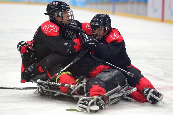 Three men on sleds lying on the ice and hugging each other in full Para ice hockey gear.