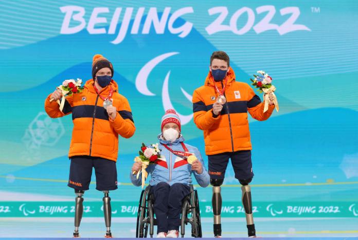 Norway's Jesper Pedersen holds up his gold medal and a bouquet of flowers during a victory ceremony at Beijing 2022 while Dutch skiers Jeroen Kampschreur and Niels de Langen stand next to him, showing their silver and bronze medals, respectively
