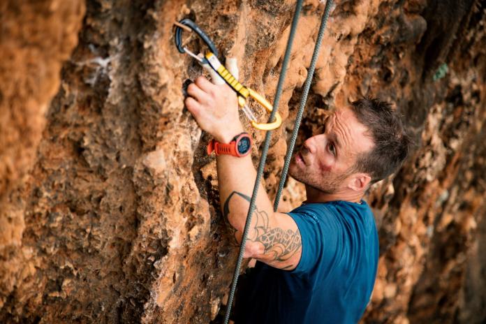 A close up of Andrea Lanfri as he scales a side of a mountain in the summer with the help of climbing equipment.