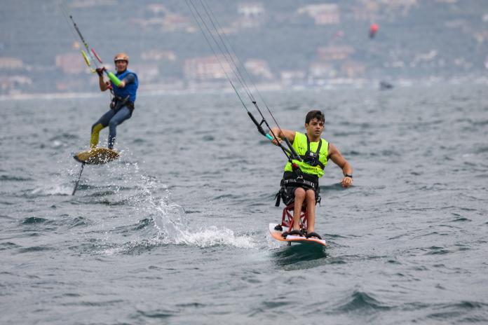 Two Para kiteboarders on the water. The one at the front is sitting on a special board and the one in the background is a standing kiteboarder with an arm prosthetic.