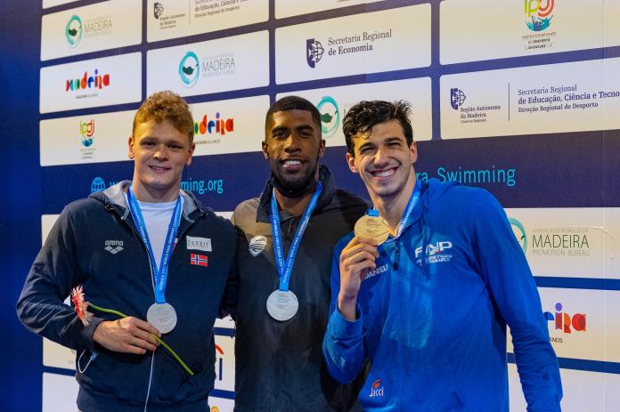 Three smiling swimmers show off their medals after a medal ceremony.