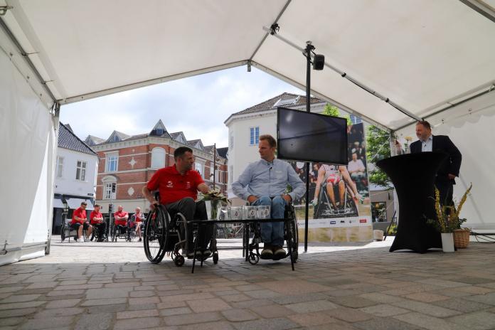 Two men in wheelchairs and one man behind the podium prepare to start the draw under a tent roof.
