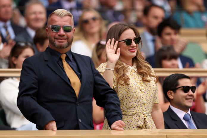 A man and a woman in smart dress smile as they are cheered by Wimbledon spectators.