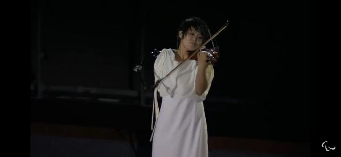 A woman in a white dress plays the violin with a bow attached to a specially designed prosthetic arm at the Opening Ceremony of the Tokyo 2020 Paralympic Games.