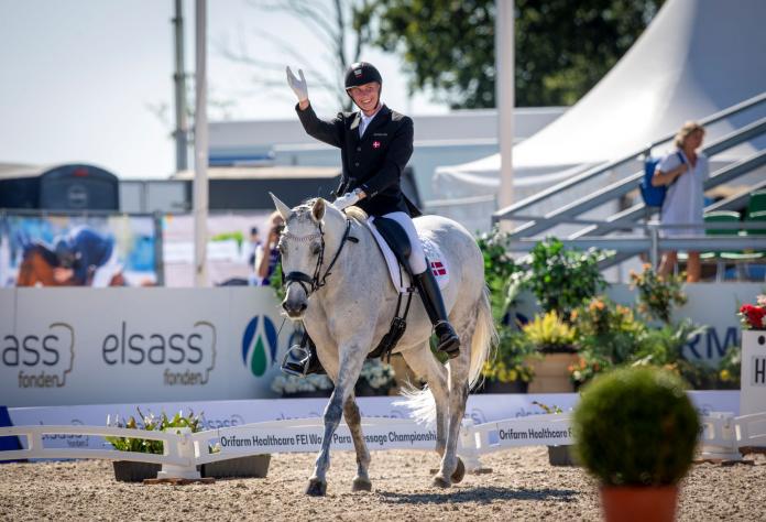 A male athlete riding a horse waves to the camera.