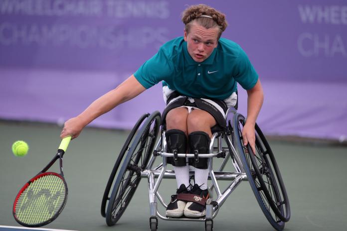 A male wheelchair tennis player returns a volley during a match on a hard court.