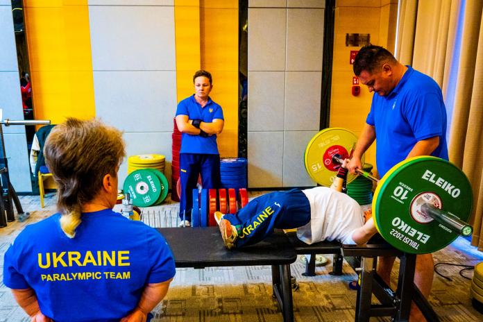 An athlete lifts a barbell in training as coaches look on.