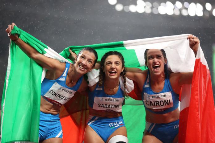 Three female athletes hold the Italian flag while placing their arms over each others' shoulders.