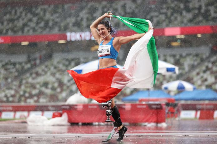 A female athlete with a prosthetic leg holds Italy's flag in the stadium.