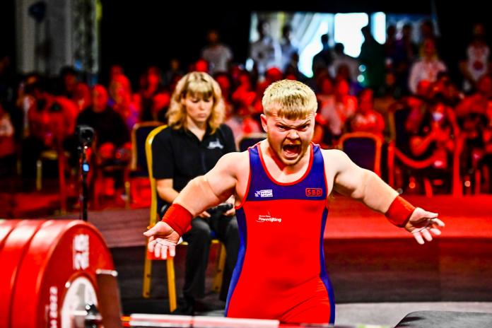 A male Para powerlifter yells in front of the barbell with his arms wide.