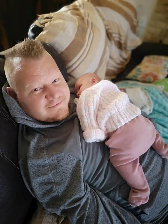 A man in a sweatshirt sits on the couch with a newborn girl sleeping on his chest.