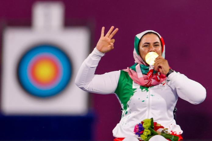 A female athlete kisses a gold medal and raises three fingers to show that she has won three Paralympic gold medals.