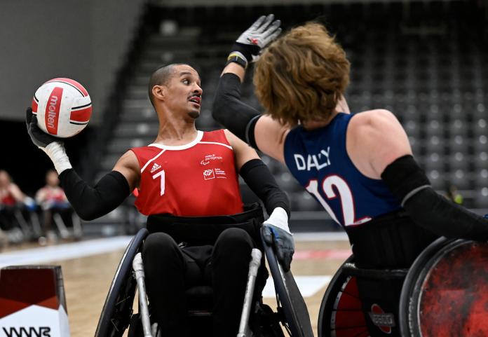 A wheelchair rugby player holds a ball with his right hand, while another player tries to block him