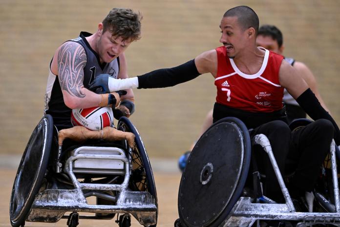 A male wheelchair rugby player reaches for a ball carried by another player.