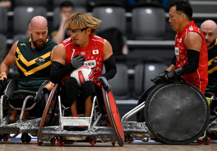 A wheelchair rugby player carries a ball, while chased by another athlete